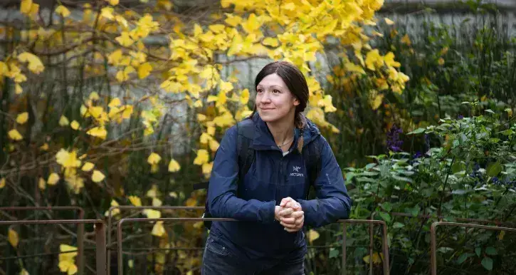 Ana Grau-Galofré souriante, en veste bleu foncé et sac à dos, s’appuie sur une barrière dans un jardin. Derrière elle, un arbre aux feuilles jaunes illumine la scène, créant une belle ambiance automnale.