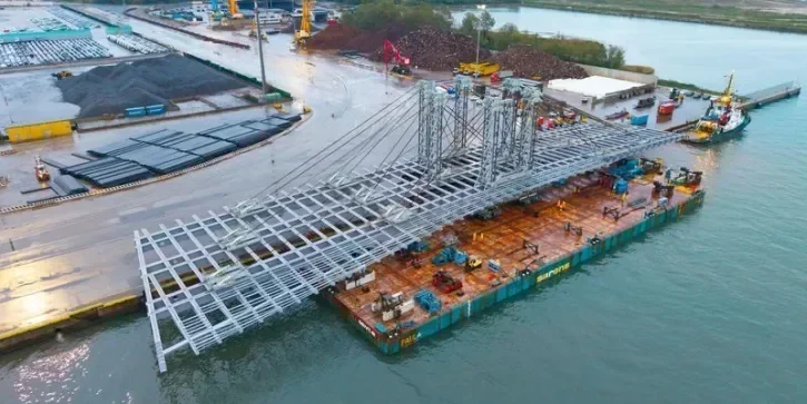 L'immense charpente métallique est chargée sur la barge à l'aide de kamags. Stationné devant sur le port, un bateau semble tout petit