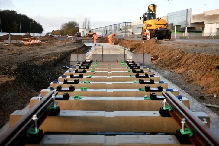 Au premier plan se trouvent les rails posés sur des traverses en béton, et en arrière-plan deux personnes en orange travaillent sur le chantier.