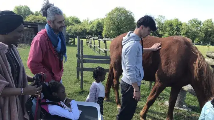 Une femme et sa fille en fauteuil roulant se rapprochent de chevaux qui sont en train d'être caressé par un jeune homme sous le regard d'un autre homme au second plan. 