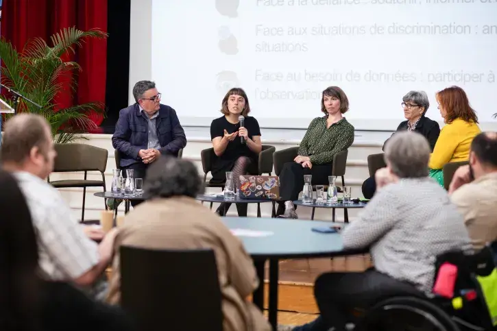 4 élus, 1 homme et deux femmes ainsi que l'animatrice et une sociologue sont assis en ligne sur la scène, sous le regard de plusieurs personnes assises à une table ronde au premier plan