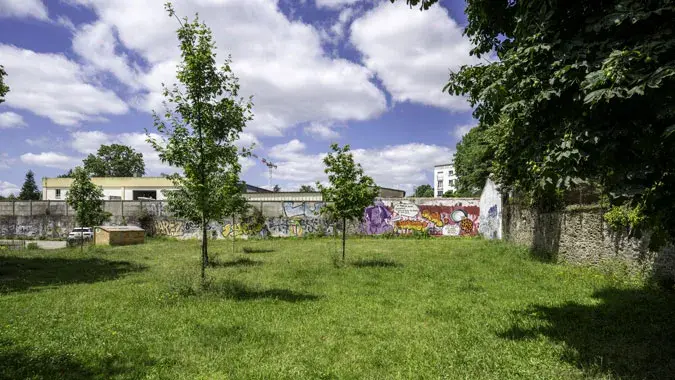 L’espace vert Simone Iff, d’une superficie conséquente d’environ 1 000m², comprend un grand arbre majestueux et de jeunes pousses récemment plantées. Il est délimité par des murs en pierre et un mur de clôture, la halle ne faisant pas partie du lieu à réi